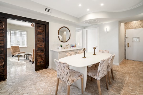 This image shows a modern dining room with a white table and wicker chairs, adjacent to a living area with chairs and a window.