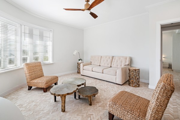 A modern living room with a beige sofa, two woven chairs, unique wooden tables, and a ceiling fan. Bright natural light from large windows.