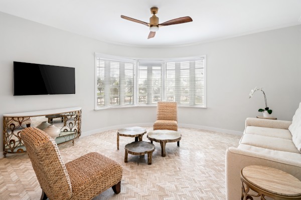 A modern living room with wicker chairs, a white sofa, small round coffee tables, a wall-mounted TV, and a ceiling fan near a bay window.