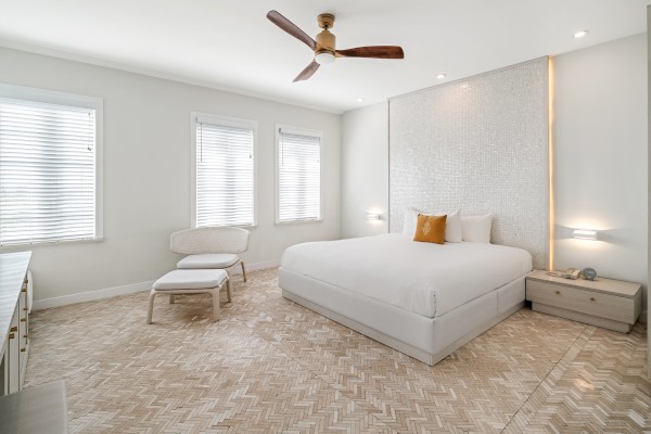 The image shows a modern bedroom with a bed, nightstands, a chair, a ceiling fan, and windows, featuring a simple and elegant design.