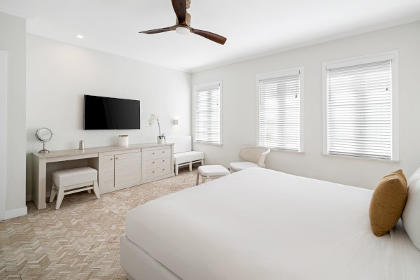 The image shows a bright bedroom with a large bed, TV, ceiling fan, dresser, two benches, and windows with blinds.