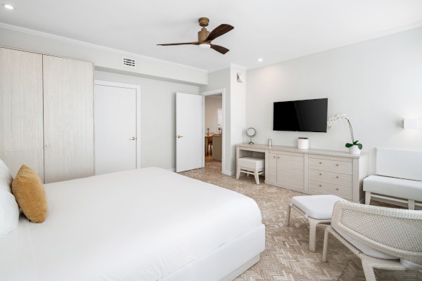 A modern, minimalist bedroom with a large bed, neutral colors, a TV on the wall, armchair, desk, and ceiling fan.