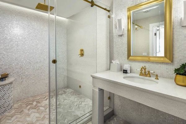 The image shows a modern bathroom with a glass shower enclosure, gold fixtures, a white countertop with a sink, and a large framed mirror on the wall.