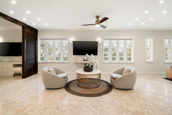 A modern living room with two gray chairs, round coffee table, TV, and ceiling fan. Large windows provide ample natural light.