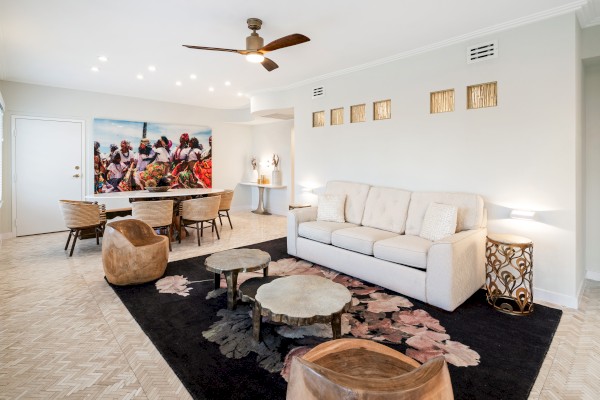 A modern living room with a beige sofa, black rug, wooden tables, a dining area, ceiling fan, and colorful wall art completes the decor.