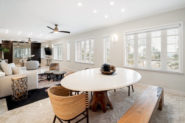 A modern living and dining area with a large table, wicker chairs, and ample natural light streaming through multiple windows.