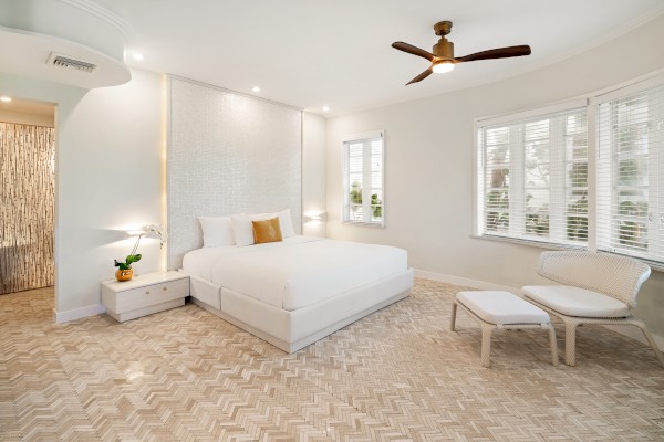 A modern bedroom with a large bed, white walls, a ceiling fan, and a seating area, featuring natural light and minimalist decor.