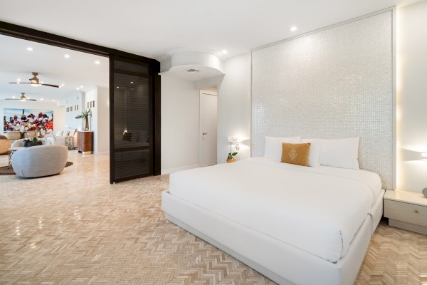 A modern bedroom with a white bed, textured wall, and open space leading to a living area with seating and decor.