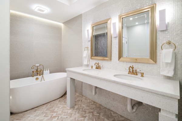 The image shows a modern bathroom with a white bathtub, dual sinks, gold fixtures, two mirrors, beige tiles, and elegant lighting fixtures.