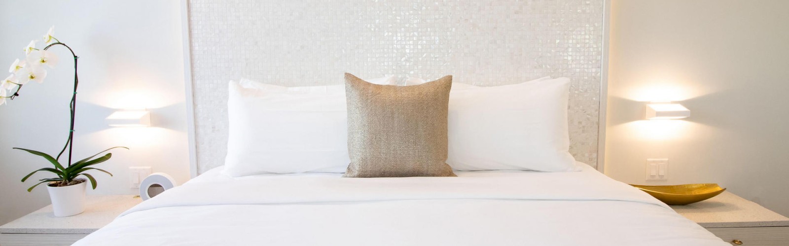 A neatly made bed with white sheets and three pillows, a beige one in the center, flanked by bedside tables, a plant, wall lights, and a clock.