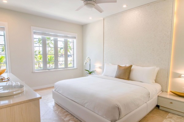 A bright bedroom with a large bed, bedside tables, a ceiling fan, and windows letting in natural light. Decor is minimalistic and elegant.