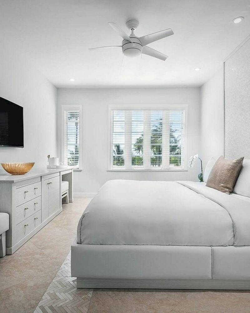 A modern bedroom features a large bed with white linens, a wall-mounted TV, a side bench, and a dresser with a decorative bowl, ending the sentence.