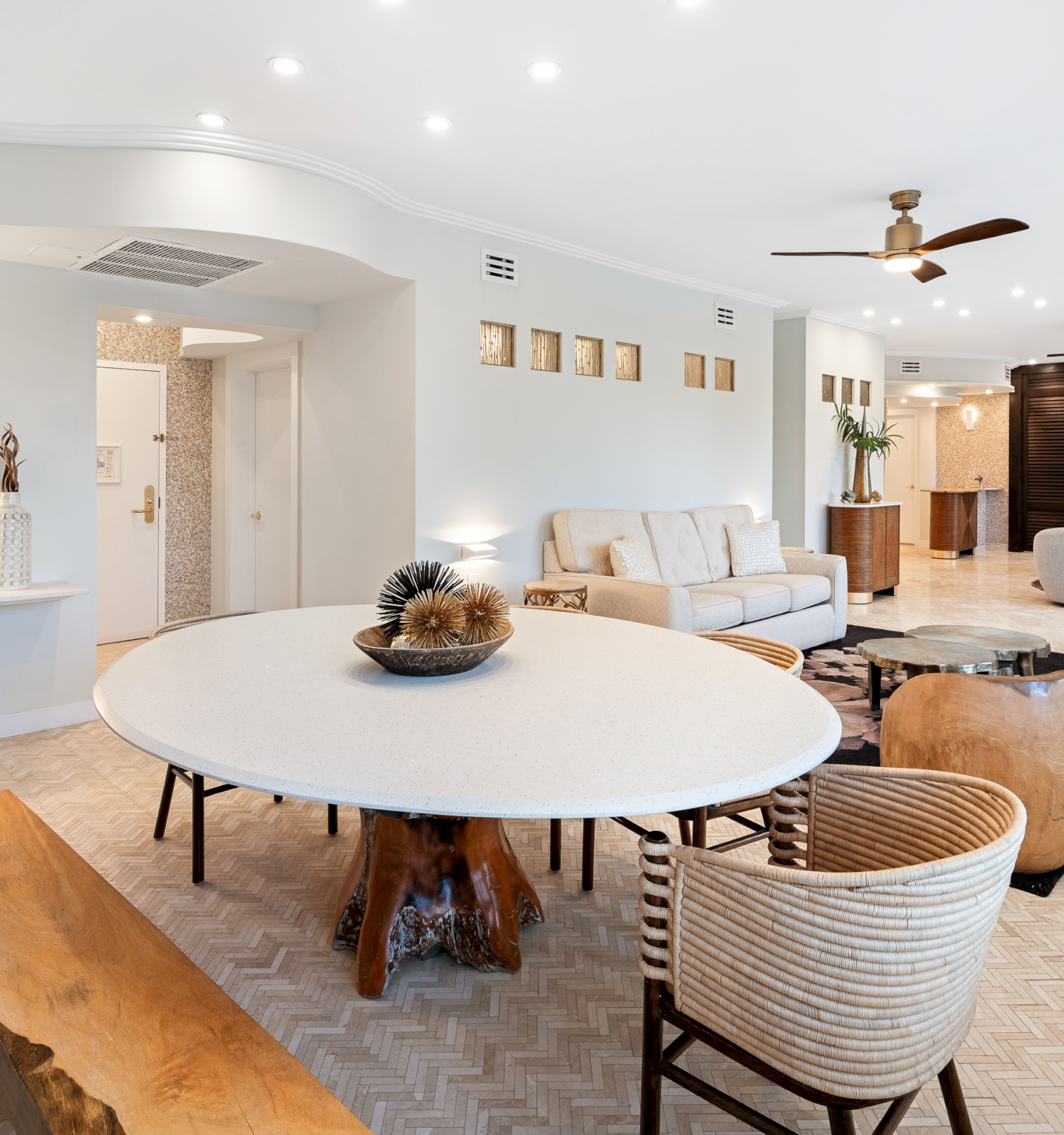 A modern, open-concept living area with a large round table, chairs, sofa, and decor. Bright lighting and ceiling fans enhance the space.