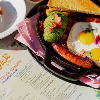 A breakfast setting with a toasted bagel sandwich, sunny-side-up eggs, avocado, salsa, sausage, and a colorful menu titled 