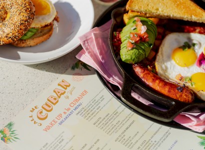 A breakfast setting with a toasted bagel sandwich, sunny-side-up eggs, avocado, salsa, sausage, and a colorful menu titled 