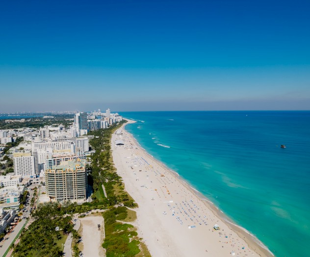 An aerial view of a coastal city with high-rise buildings, a long sandy beach, and clear blue waters extending to the horizon.