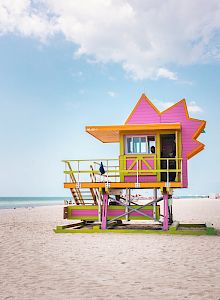 A colorful lifeguard tower sits on a sandy beach, with a few people in the distance and the calm ocean under a partly cloudy sky.