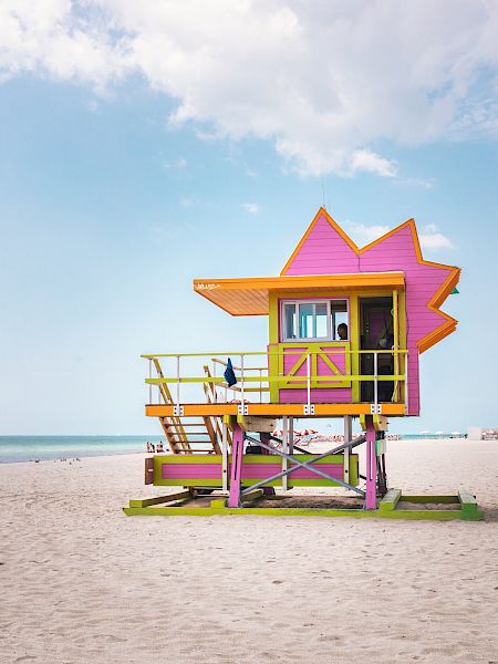 A colorful lifeguard tower sits on a sandy beach, with a few people in the distance and the calm ocean under a partly cloudy sky.