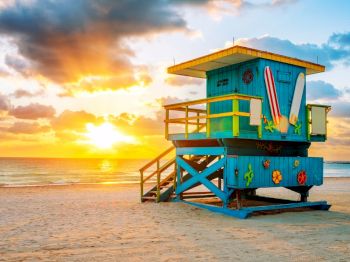 A colorful lifeguard tower is on a sandy beach at sunrise, with vibrant clouds in the sky and the sun reflecting off the calm ocean water.