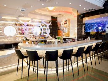 A modern bar with a sleek counter, black bar stools, a well-stocked shelf, soft lighting, and a large screen on the wall.