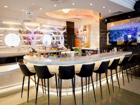 A modern bar with a sleek counter, black bar stools, a well-stocked shelf, soft lighting, and a large screen on the wall.