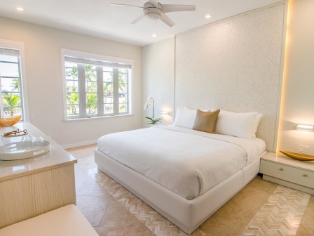 A bright, modern bedroom with a large bed, white bedding, a ceiling fan, and a window with a view of greenery. There is a dresser and two nightstands.