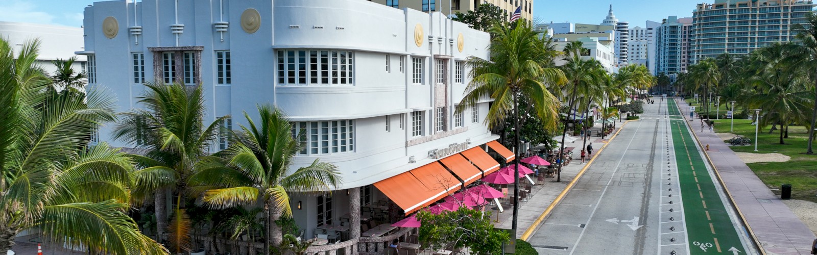 Street view of a coastal city with a historic building, palm trees, bike lane, and outdoor seating under red umbrellas.