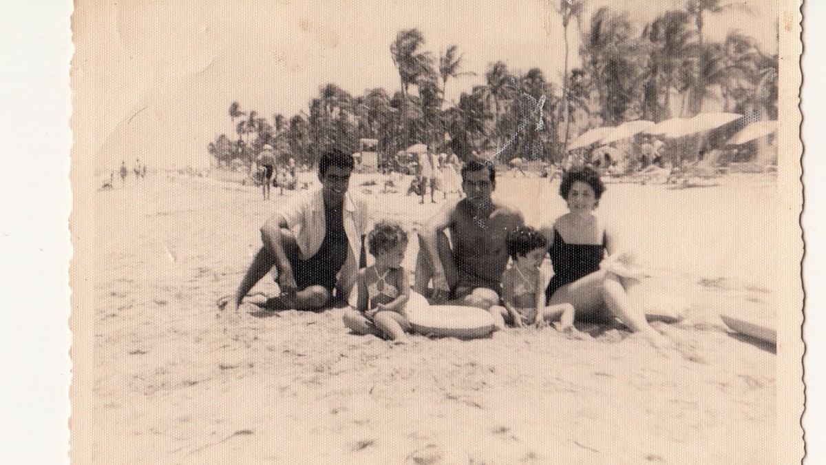 A vintage photo of five people sitting on a sandy beach, surrounded by palm trees, with additional beachgoers and umbrellas in the background.