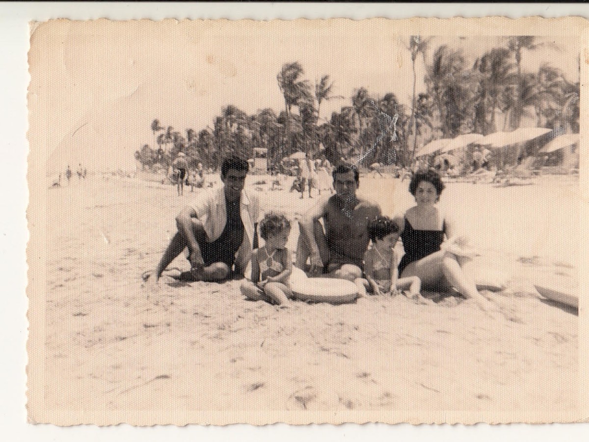 A vintage photo of five people sitting on a sandy beach, surrounded by palm trees, with additional beachgoers and umbrellas in the background.