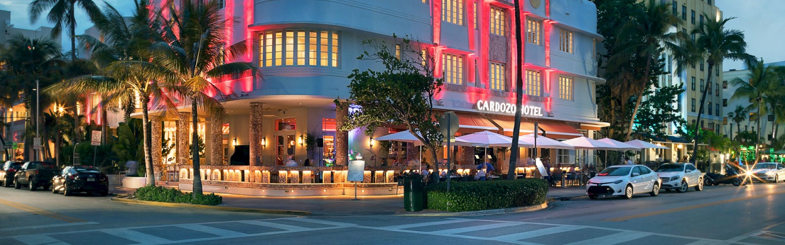 An art deco building with bright pink lights on a busy street corner during dusk, with palm trees and cars.