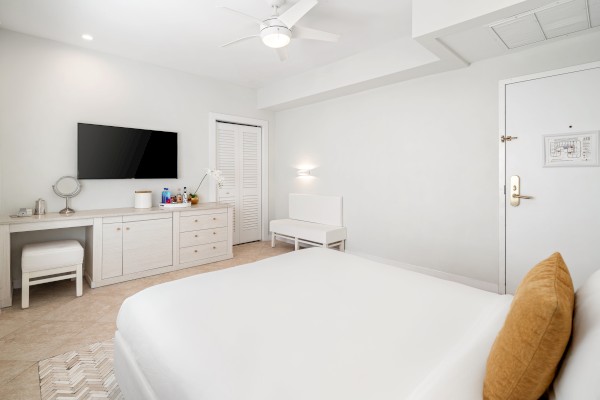 A modern, minimalist bedroom with a white bed, TV, desk, and chair. Walls are light-colored, and there’s a ceiling fan and closed door.