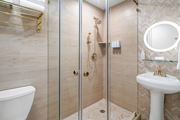 A clean bathroom features a glass shower, pedestal sink, toilet, and gold fixtures. The walls have a light wood texture, adding elegance.