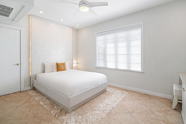 A minimalist bedroom with a white bed, two pillows, a patterned rug, and a window with blinds, plus a ceiling fan and a corner desk.