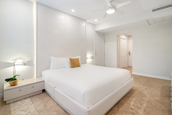 A modern bedroom with a large white bed, side table with a plant, ceiling fan, and soft lighting, creating a minimalist and cozy ambiance.