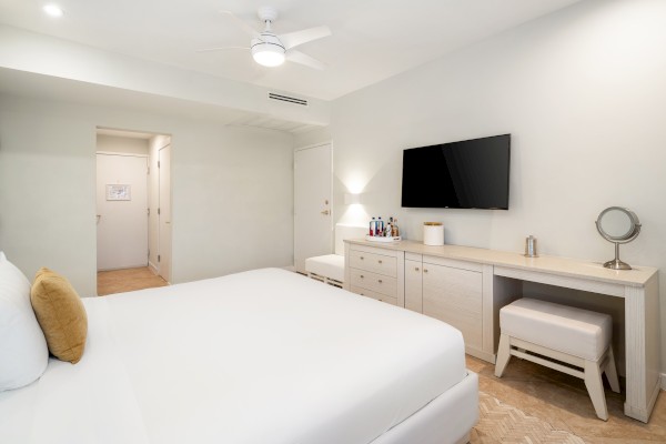 A modern bedroom with a bed, TV, dresser, and mirror. The room has white walls and a ceiling fan.