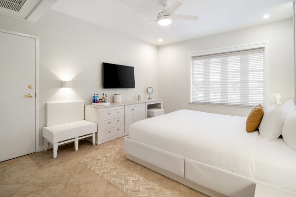 A modern bedroom with a large bed, wall-mounted TV, dresser, chair, and window with blinds. Bright and minimalistic design.