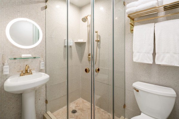 The image shows a small bathroom with a glass shower, round mirror, sink, and toilet. Towels are neatly stacked on a gold rack.