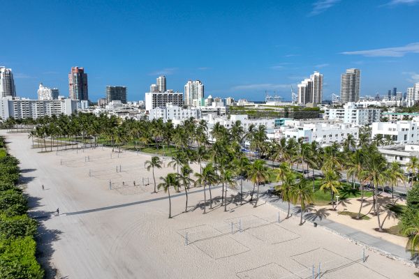 An aerial view of a beach with volleyball courts, palm trees, and a cityscape with numerous high-rise buildings under a clear blue sky.