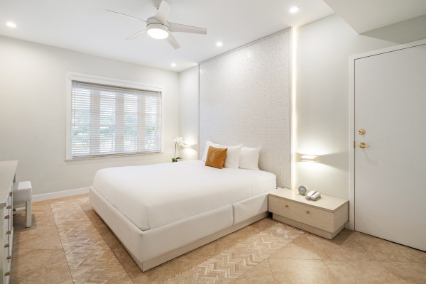 A modern bedroom with a double bed, ceiling fan, side tables, and a window with blinds. Neutral colors create a calm atmosphere.