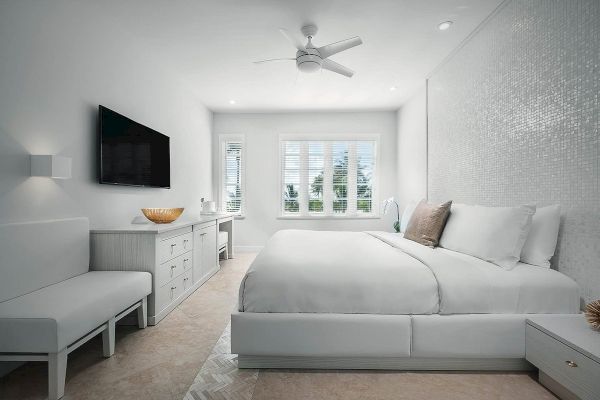 A modern bedroom with a white theme, featuring a bed, TV, dresser, seating area, and ceiling fan, illuminated by natural light from large windows.