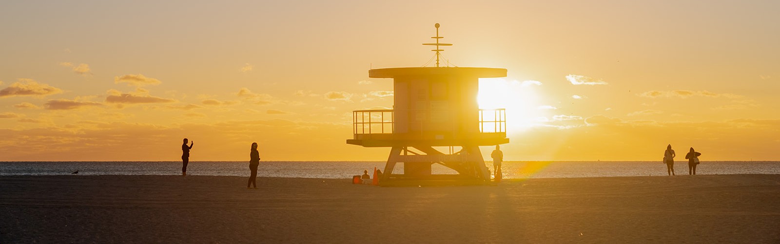 A lifeguard tower on a beach at sunset, with a few people standing and walking around, casting elongated shadows on the sand.