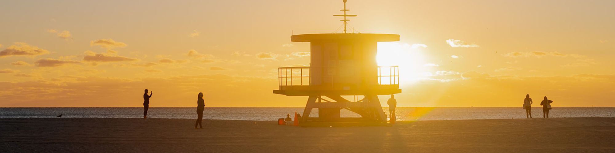 A lifeguard tower on a beach at sunset, with a few people standing and walking around, casting elongated shadows on the sand.