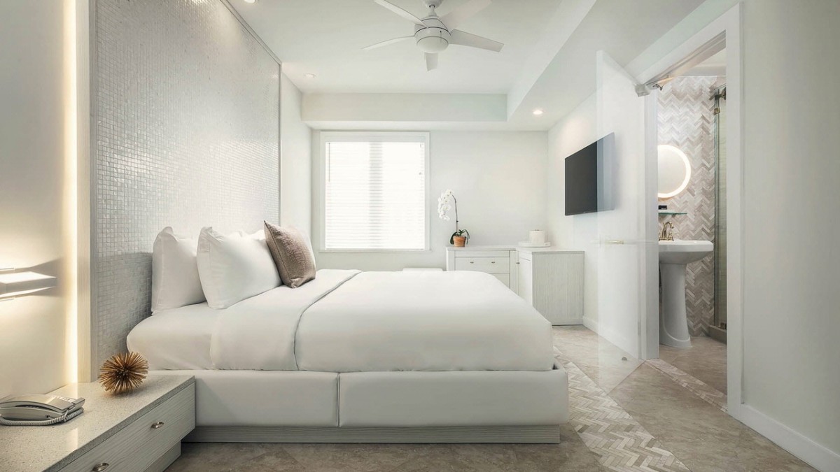 A modern bedroom with a large bed, minimalist decor, and neutral tones; it features a flat-screen TV mounted on the wall and an ensuite bathroom.