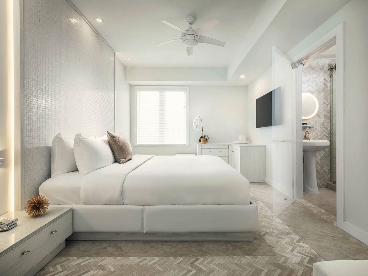 A modern bedroom with a large bed, minimalist decor, and neutral tones; it features a flat-screen TV mounted on the wall and an ensuite bathroom.