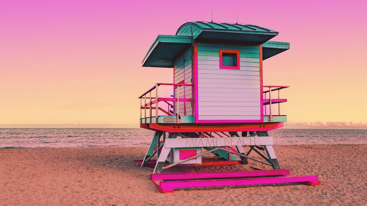 A colorful lifeguard tower with pink, teal, and white accents stands on the beach under a vibrant pink and yellow sky.