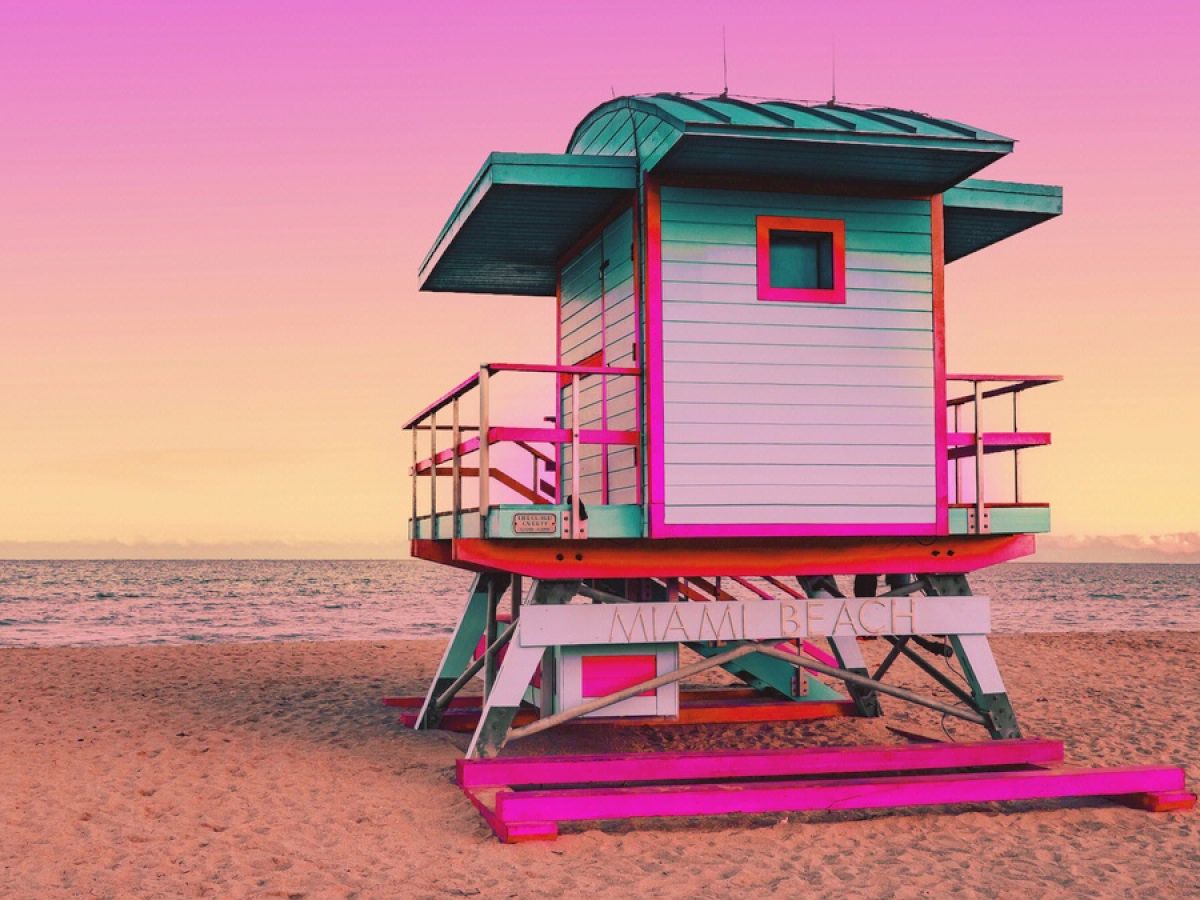 A colorful lifeguard tower with pink, teal, and white accents stands on the beach under a vibrant pink and yellow sky.