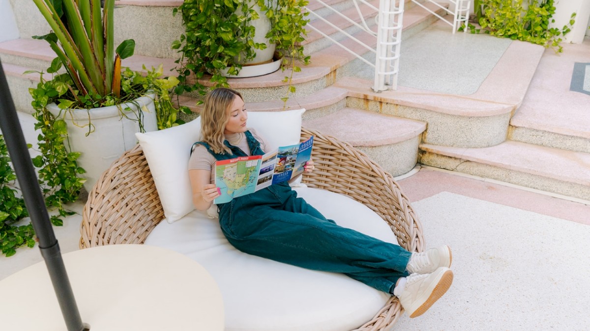 A woman relaxes on a round wicker lounge chair, reading a map or magazine, surrounded by lush greenery and steps in an outdoor setting.