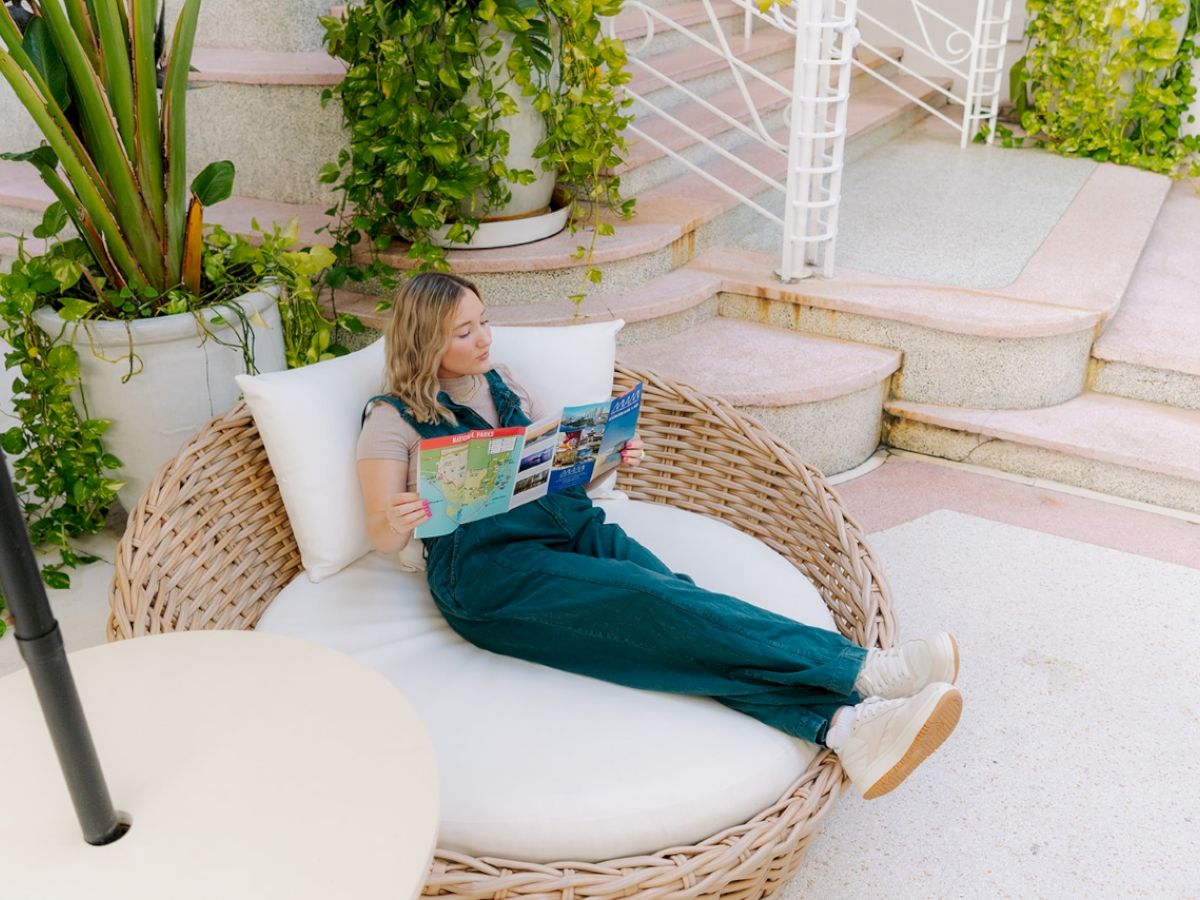 A woman relaxes on a round wicker lounge chair, reading a map or magazine, surrounded by lush greenery and steps in an outdoor setting.