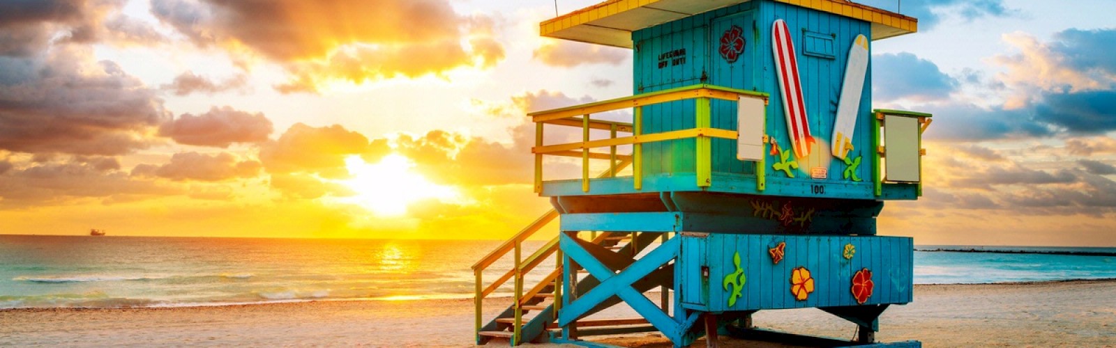 A colorful lifeguard tower stands on the beach with a sunrise, surfboards, and ocean waves in the background, under a partly cloudy sky.