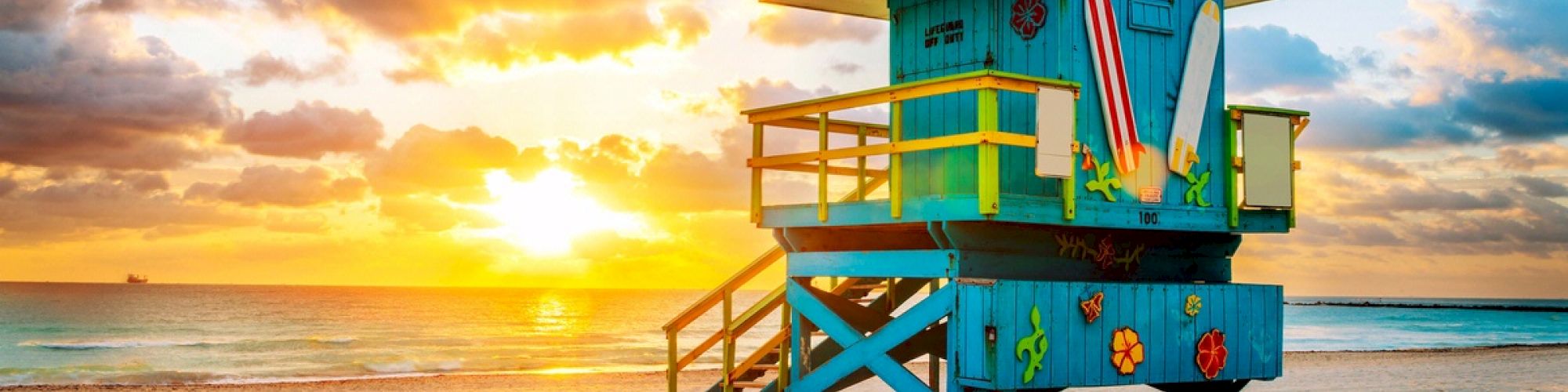 A colorful lifeguard tower stands on the beach with a sunrise, surfboards, and ocean waves in the background, under a partly cloudy sky.